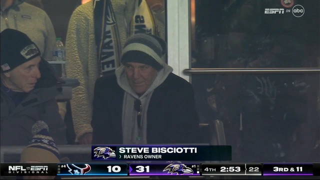 A man in winter attire (Steve Bisciotti, Baltimore Ravens owner) watching a sporting event from a glass-enclosed area with an on-screen caption indicating his association with a sports team. The scoreboard displays a late-game situation.