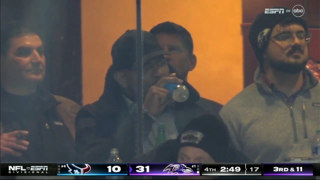 Spectators in a suite (in the middle is Ray Lewis drinking a bottle of water) watching an American football game, with on-screen graphics showing the score, quarter, time left, and down.