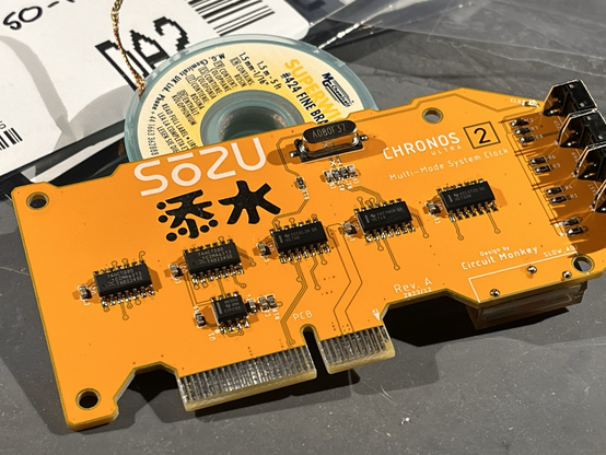 Close up of the Sozu clock card. There is a row of surface mount chips soldered along the centerline of the board. There are push switches to the right, on the boards front edge. There is a 8MHz crystal soldered at the top center.