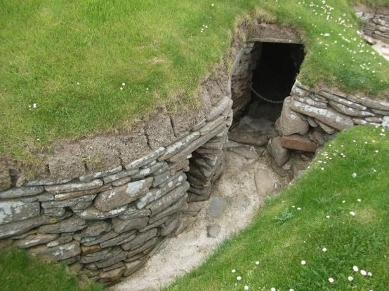 Photo is of a grassy daisy-covered bank and an uncovered neolithic corridor between houses at the settlement at Skara Brae, in the Orkney islands.  the corridor consists of layers of flat, carefully shaped rock that form curving walls with niches cut out for doorways, and the floor is sandy with a few stepping stones showing. Caption adds, I can imagine children running and playing in these corridors.