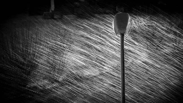 Schwarzweissbild einer Laterne, in derem Licht ein Schneegestöber als feine verwirbelte Linien zu erkennen ist.