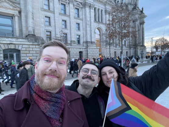 Three people outside the German Bundestag with other protestors in the background. A progress flag is in the foreground.