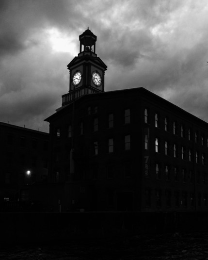 Black and white photo of a silhouetted mill building that features a clock tower. There is one window lit from within, and one exterior light visible. There are threatening clouds in the sky behind it. The clock reads 4:44.