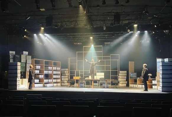 Stage with actors, a table, chairs, and stacks of file boxes under dramatic lighting.