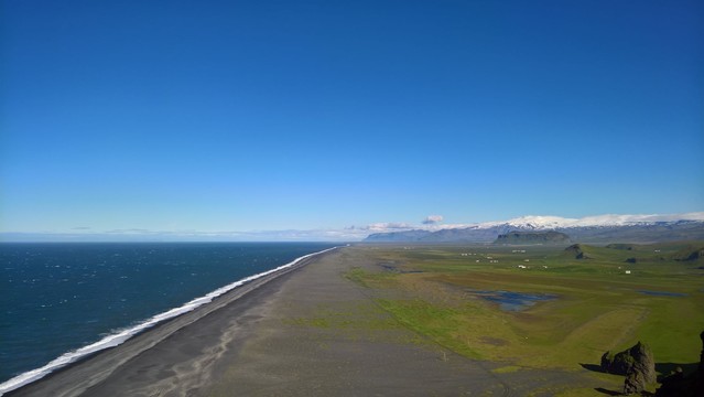 Man sieht die Wasserlinie bis zum Horizont. Links ist der Atlantik in einem tollen Blau, in der Mitte der dunkle, teils schwarze Strand, rechts eine große, grüne Ebene, in der Ferne Berge mit Gletscher. Der Himmel ist wolkenfrei.