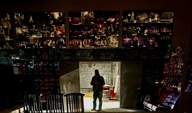 Interior of a dimly lit room with photographic artwork on the wall, a person standing near a doorway, and shelves with various items reflected in a mirror.