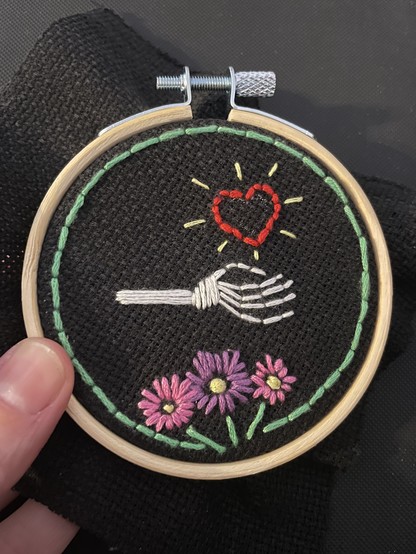 A hand holding a small round embroidery hoop covered with black fabric. Embroidered on is a skeleton hand with a red heart with sunbeams floating above its palm. Below the hand are 3 flowers embroidered in purple and lilac. A green line is embroidered around the hoop.