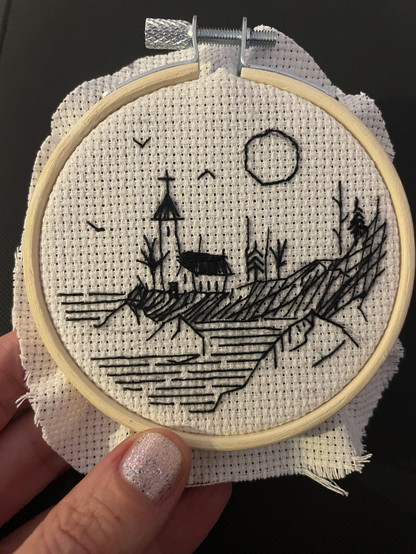 A hand holding a small round embroidery hoop covered with white farbric. A mountainous beach landscape with a small church and a few trees are embroidered in black thread. A round sun stands above the landscape, with three small birds flying next to it. The embroidery looks as if it has been roughly drawn with black fineliner.