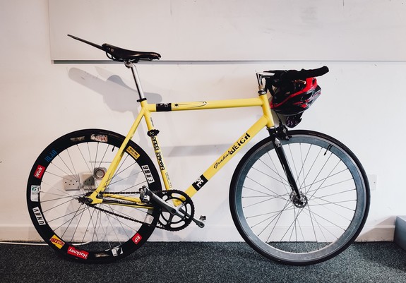 A yellow single speed bike with bullhorn track handlebars leans against a white wall. The rear wheel has numerous stickers on the deep dish of the rim