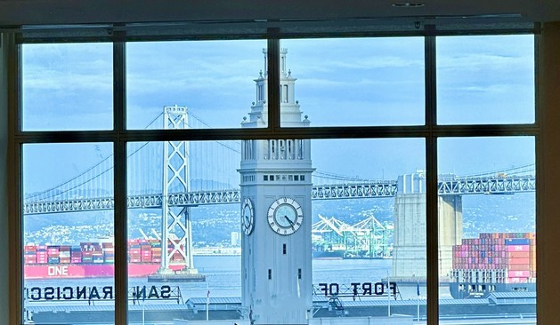 View out a window of the San Francisco Bay. The San Francisco ferry building clock tower is in the foreground, the Bay bridge is in the background, and there are two ships one inbound and one outbound passing under the bridge.
