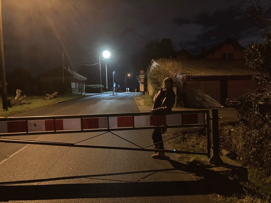 A night photo with light from two street lampposts. The author is resting on a red and white metal barrier stretched across the road, locked shut to stop cars. Rest assured: other places are open to cross at night, further along!