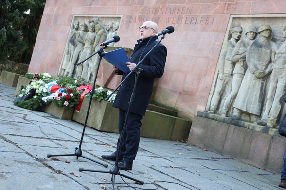 Oberbürgermeister Sven Schulze steht am Mikrofon und hält eine Rede. Im Hintergrund ist das Mahnmal im Park der Opfer des Nationalsozialismus zu sehen. Davor liegen Kränze, die zuvor niedergelegt wurden.