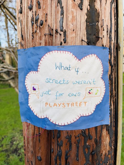 An embroidered cloth attached to a telegraphy pole. The stitching says: what if… streets weren’t just for cars. Playstreet