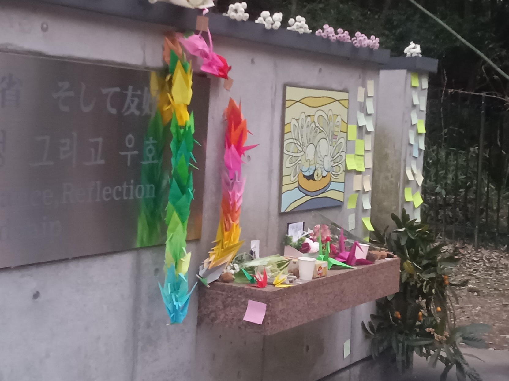 追悼碑正面、飾られる千羽鶴やお供え物、付箋。平和の鳩、『記憶 反省 そして友好』