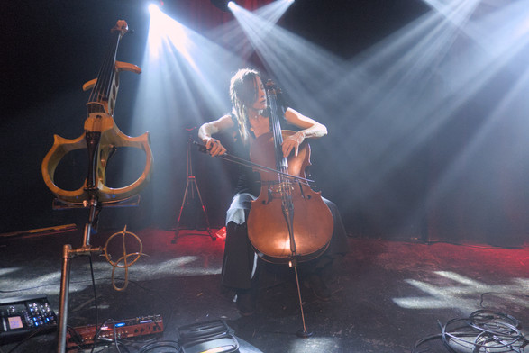 Jo Quail playing cello on stage. Her eyes are closed as she concentrates intently on the music A skeletal electric cello is on its stand to her side.
