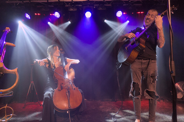 Jo Quail sat playing cello alongside Jon Gomm stood playing acoustic guitar on stage. They are both smiling broadly in enjoyment at each other's playing.