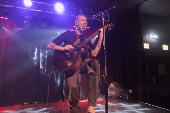 Jon Gomm playing acoustic guitar on stage. He has a wide smile and is stood with one leg raised forwards.