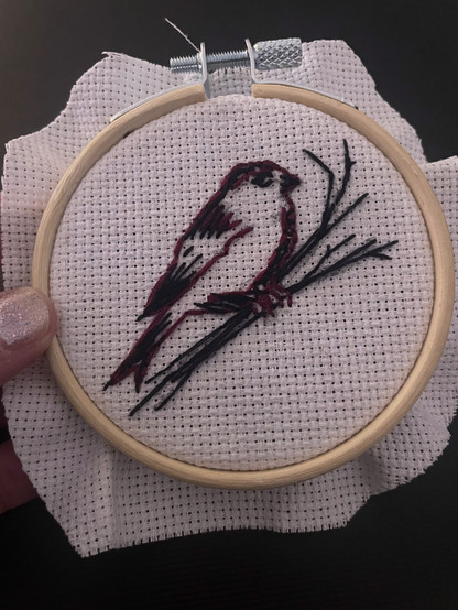 A hand holds a small, round embroidery frame, covered with white embroidery fabric. On it is a small bird on a branch, embroidered in the colours red and black.