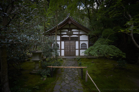 A small, white-panelled Japanese building, with pitched concave-slope roof, clad in convex bamboo-resembling tiles. The building is at the centre of frame, enclosed on all sides by lush, dark green trees and shrubs. The ground in front is extensively covered in bright green moss, and down the centre runs a flagstone path, with a bamboo barrier preventing access.