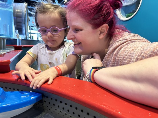 Ripley with purple glasses and messy hair playing in the water at a children’s museum with my wife who has pink hair