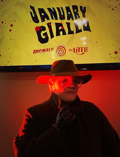 Meghan in a hat and trench coat, red light on the side of her face, with a screen above that reads "January Giallo."