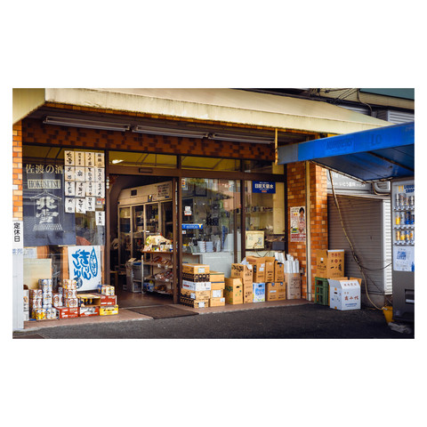 Captured in the photo is a local liquor store, a staple in the neighborhood for quick pick-ups of convenience food and various snacks. The storefront is a vibrant tableau of everyday life, offering a glimpse into the community's routine, with items catering to both necessity and pleasure neatly displayed in its welcoming interior.