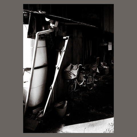 In this monochromatic composition, a bicycle equipped with cloth-covered baskets takes refuge in the shade, providing a stark contrast to the sunlit day. It rests beside a home whose better days are recounted through the visible wear and tear, the structure a patchwork of repairs and resilience, evoking the wabi-sabi aesthetic of beauty in imperfection.