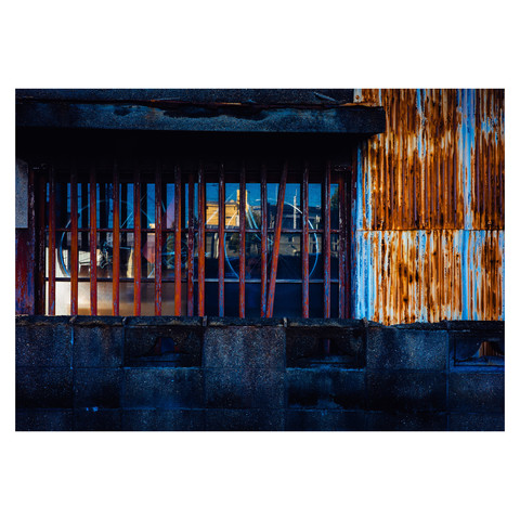 Through the barred window of a garage, the silhouette of a bicycle hangs aloft, adding a touch of the familiar to a canvas of rust and wear. The bike, perched above the ground as if on display, hints at stories of travel and rest, of motion and pause, within the confines of its urban frame.