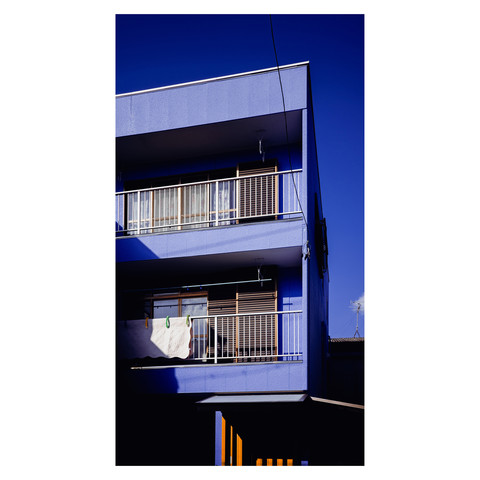 The striking purple facade of an apartment building pops against the backdrop of a clear blue sky. The saturated colors create a visual feast reminiscent of a refreshing glass of grape juice, while a solitary towel hangs on the balcony, basking in the vibrant glow of a sunny day. The building, a bold statement in an urban landscape, stands out as a testament to the city's playful character and vivid life.