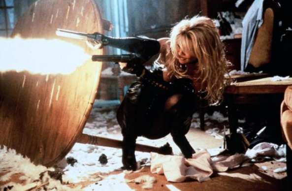 an image from the movie, pamela anderson as barb wire in the 1996 film, crouching behind an overturned table, shooting two pistols, fire spewing out of the one in her left hand 