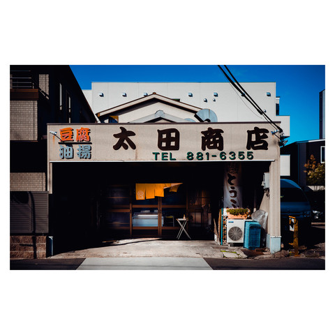 The photograph displays a tofu shop's charming entrance, emblazoned with traditional signage, offering a glimpse into a space where craftsmanship and community intersect. The sun casts deep shadows, highlighting a scene that invites passersby to step in and savor the simplicity and comfort of a neighborhood staple.