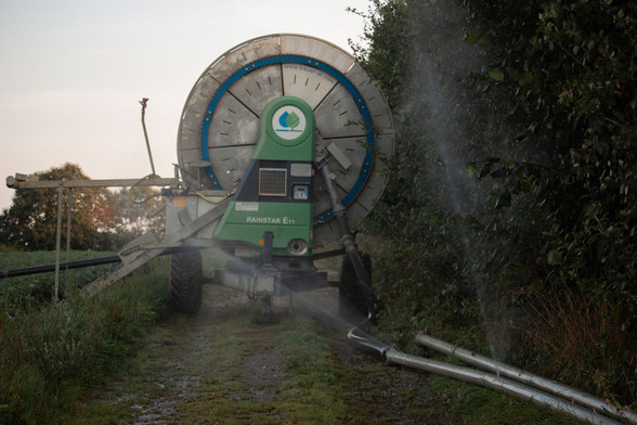 Vue frontale de l'arrière d'un imposant enrouleur d'irrigation planté au beau milieu d'un chemin, empêchant tout passage. A droite, une haie et des tuyaux perforés d'où jaillit des gerbes d'eau, à gauche, le champ et le tuyau d'irrigation.