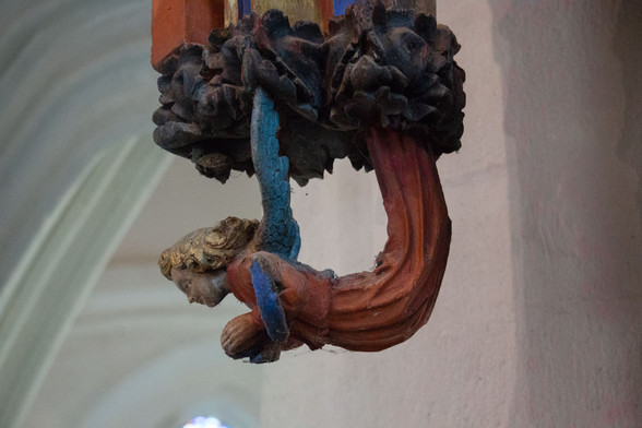 Détail du jubé en bois d'une chapelle de cornouaille morbihannaise. Un angelot au costume rouge et bleu a les pieds rattachés au bas d'un montant ouvragé. Il se redresse pour regarder, mains jointes, l'assistance potentielle.