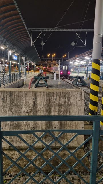 Picture of an empty part of a train station in the dark. There are two platforms surrounded by tracks from both sides.
