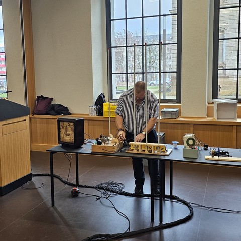Carsten Hörner baut Technik im IdeenReich der Universitätsbibliothek Chemnitz für seinen Experimentalvortrag zum Audion auf. Auf einem Tisch sind dazu einige technische Geräte nebst Kabeln zu sehen.