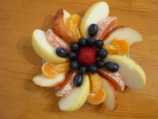 Fruit plate. 4 of the 8 slices of a Nashi pear, 4 of the 8 slices of a red pear, all arranged like petals around a strawberry in the middle. There are 4 blood orange slices and 8 small mandarin slices, as well as black grapes around the central strawberry.