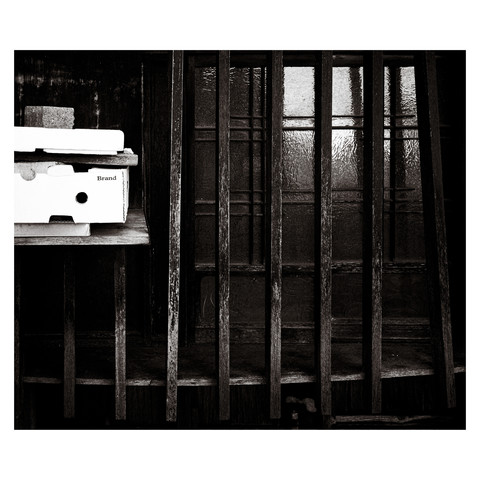 The monochromatic image captures a moment in time at the traditional-style window of an old house, the textured glass slightly obscuring the world beyond. A lone shelf holds a produce box marked with a single enigmatic word: "brand." It stands as a symbol, a minimalist representation of identity and history, set against the backdrop of a window that has borne witness to the ebb and flow of countless days.