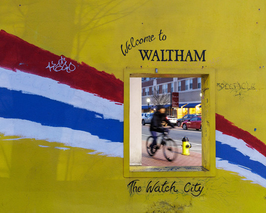 Color photograph of a wall alongside a city sidewalk and street. The wall has a medium size “window” in it where the viewer can look through to see the street and sidewalk at an angle. The wall is painted yellow with four adjacent red, white, and blue stripes painted diagonally across it in an arc—the window bisects this band of color. There is writing above window, “Welcome to WALTHAM.” Just below that (and still above the window) is scrawled, “THE COPS SUCK.” Below the window is written, “The Watch City,” Waltham’s motto. To the right of the window is written, “SPECTACLE,” and that word is partially obscured by a series of X’s written over it. Through the window, the viewer can see a slightly blurred person riding a bicycle on the brick sidewalk past a fire hydrant, some parked cars, one moving car, and a building across the street beyond.