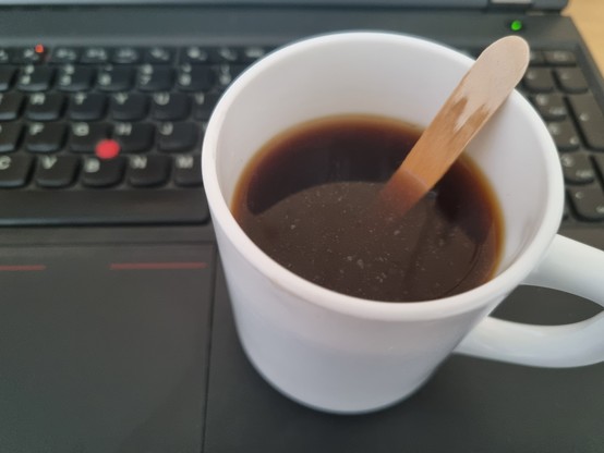 Ein Becher Krankenhauskaffee mit hölzernem Einweglöffel, der mir heute aus unerfindlichen Gründen reingetan wurde. Es ist nur ein Tropfen Milch im Kaffee und kein Zucker, dafür aber ein befremdliches Holzaroma.