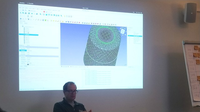 Julian sat in front of a projector. FreeCAD is projected and we can see a complex mesh showing carbon fibre wrap over a 3D object. 