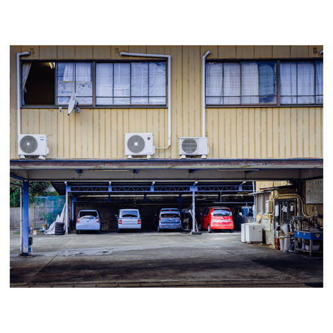 A scene of daily life is captured as vehicles rest in an open garage space, each in its designated spot. Above, air conditioning units are installed on the side of the building, suggesting a blend of functionality and domestic routine. The pale yellow walls and symmetrical windows add to the orderly aesthetic, creating a tableau of stillness in the urban day-to-day.
