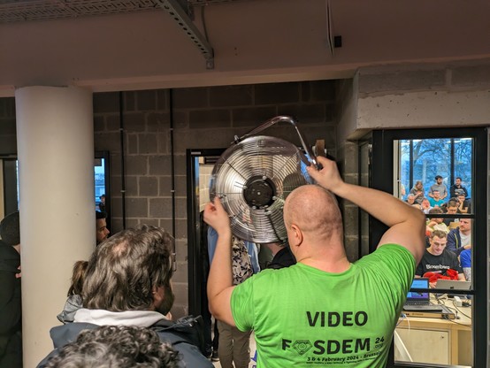A Fosdem volunteer holding an electric fan, pushing fresh air into the devroom while people switch rooms