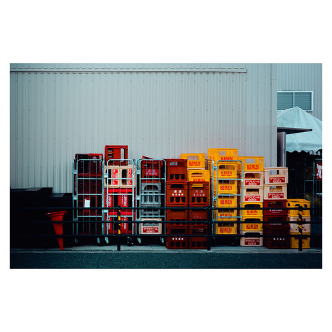 The vibrant array of beer crates creates a striking contrast against the muted backdrop of a corrugated wall. The orderly stacks sit in repose, their work of carrying frothy delights done for the day, now enjoying a moment of rest under the mellow sun. The scene captures the essence of daily life's ebb and flow, the pause between the bustling moments of demand and supply.