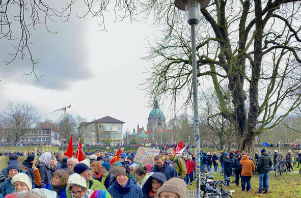 Hunderte Menschen strömen in einer Schlange mit Plakaten und Fahnen in den Händen entlang eines Weges. Im Hintergrund Gebäude wie das neue Rathaus von Hannover mit seinem grünen Kuppeldach.