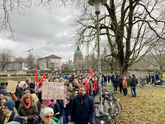 Hunderte Menschen strömen in einer Schlange mit Plakaten und Fahnen in den Händen entlang eines Weges. Im Hintergrund Gebäude .