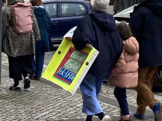 Menschen gehen einen gepflasterten Fußweg entlang. Eine ältere Person hält ein Kind an der einen Hand. Die ältere Person hat eine Art Schild unter dem Arm auf der anderen Seite. Es ist in neongelb mit bunter Schrift. Auf dem Schild steht auf grünem Untergrund Linerté, Egalité. Darunter auf blauem Untergrund steht: FckAFDé
