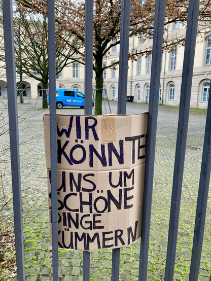 Foto mit einem Stahlzaun im Vordergrund. In dem Zaun steckt ein Pappschild mit schwarzer Schrift. Auf dem Schild steht in Großbuchstaben: Wir könnten uns um schöne Dinge kümmern. Im Hintergrund ein Parkplatz im Hof des niedersächsischen Landtags. Demonstrativ steht gut sichtbar ein blauer Kleinbus mit einer Deitschlandflagge und weißer Schrift. Auf der Seite des Autos steht: AFD.
