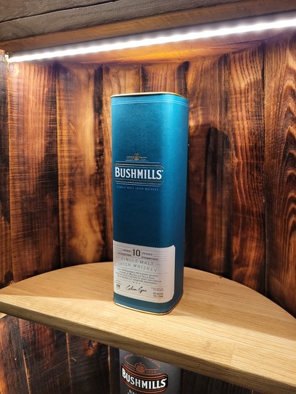 This image depicts a setting indoors, focusing on a blue cylindrical container placed on a wooden shelf. The container appears to be a bottle, likely of whiskey, given the text visible on it. The brand "Bushmills" and the words "Single Malt Irish Whiskey" can be discerned. The bottle is primarily blue with white text, giving it a distinctive appearance. The setting itself conveys a rustic charm, with warm brown hues dominating the image due to the wooden components, including the shelf and poss…