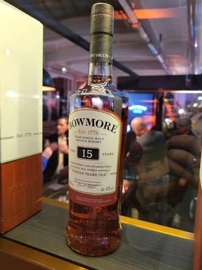 This image features a bottle of liquor, specifically Bowmore Islay Single Malt Scotch Whisky, which has been aged for 15 years, as indicated by the label. The bottle is placed on a shelf, suggesting an indoor setting, potentially a bar or a retail shop. The bottle is made of glass and appears to be full. It's noteworthy that the dominant foreground color is black, while the background is predominantly brown, likely due to the wood of the shelf. There's also an indication of a person's presence,…