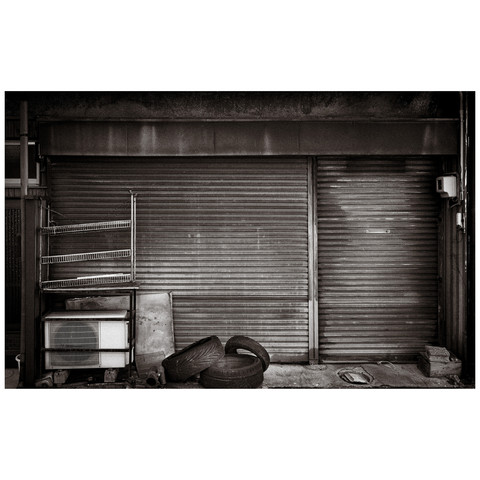 In this monochromatic scene, a collection of urban artifacts rests against a backdrop of industrial beauty. The air conditioner, silent and watchful; the racks, empty yet standing ready; the tires, echoes of journeys completed; all guarded by a shutter whose rust is the badge of its resilience and history. Together, they compose a symphony of the city's quieter moments, a narrative in texture and time.